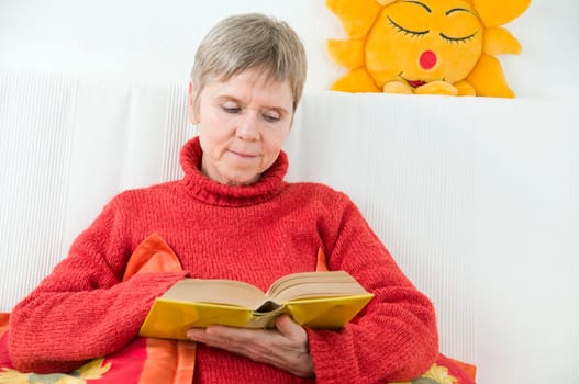 mature woman in red sweater,short and gray hair sitting on a white couch and reading a yellow book