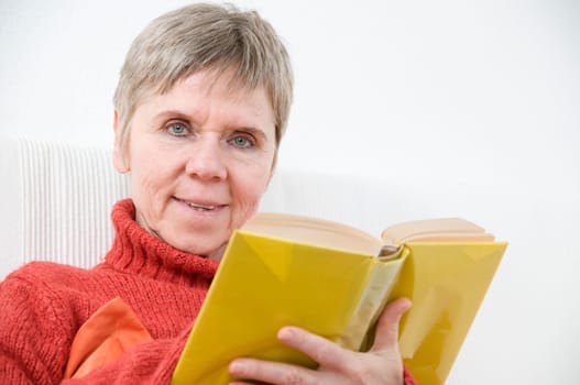 mature woman in red sweater,short and gray hair sitting on a white couch reading a yellow book