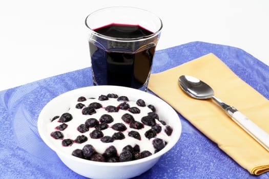 Glass of organic blueberry juice, frozen blueberries with yogurt in a bowl near a spoon on a placemat.