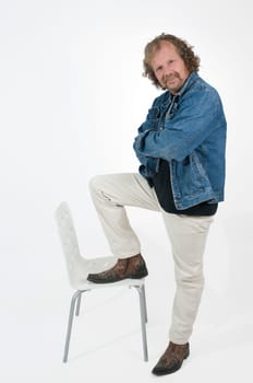 Elderly man in white pants and blue denim jacket puts one leg on the seat of a white chair