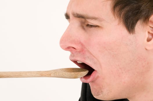 Side head shot of a young 20-year-old man with wooden spoon pointing towards his mouth and disgusted/disgusted facial expression