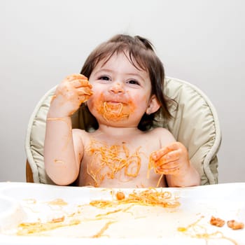 Happy smiling baby having fun eating messy covered in Spaghetti Angel Hair Pasta red marinara tomato sauce.