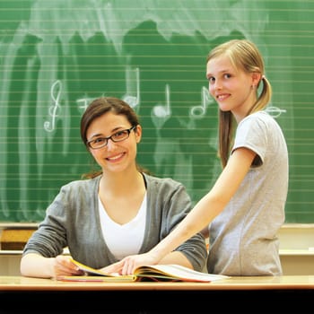 Young friendly teacher and pupil in the classroom. The teacher sits at the desk in front of the blackboard. Next to her is the student  pointing to a book  both smiling.