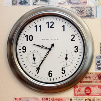 A large round atomic clock with hygrometer and thermometer on a wall with parts of a variety of money.