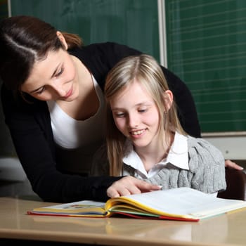 Teacher and pupil in classroom learning together. The girl looks at a book together with the teacher. She explains the task to the child. -square