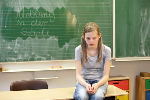 A girl sits sadly in the classroom at school in front of a blackboard with the words " Bullying at school """