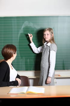 Schoolgirl in the classroom writes on the blackboard. The teacher sits at the desk and watches