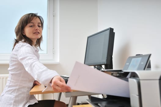 Business person sitting at table taking document from printer - monitor and workplace in background