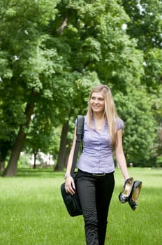 Young business woman wit laptob bag walking outdoors in park and holding shoes