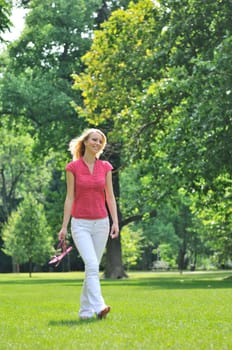 Smiling young woman holding her shoes and walking barefoot on grass in park