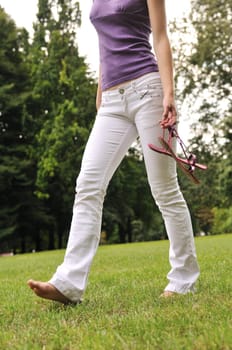 Woman (torso) walking barefoot on grass in park and holding her shoes in hand