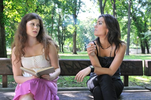 Two young people outdoors on bench in park, one teenager smoking cigarette annoys another girl