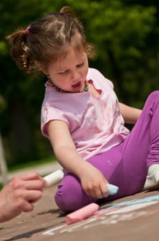 Small cute girl drawing with chalk on sidewalk, trees in background