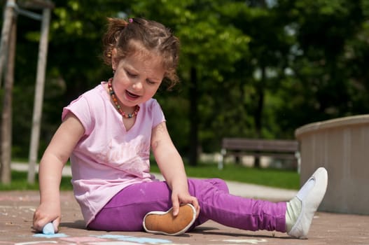 Small cute girl drawing with chalk on sidewalk, trees in background