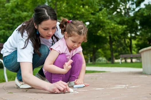 Mother is teaching her cute daughter to draw with chalk on sidewalk