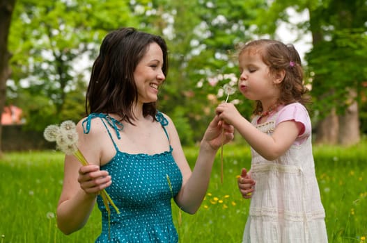 Small girl blowing to dandelion held by happy mother
