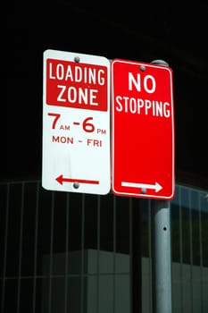 two red parking signs: LOADING ZONE and NO STOPPING, dark background, more text on signs