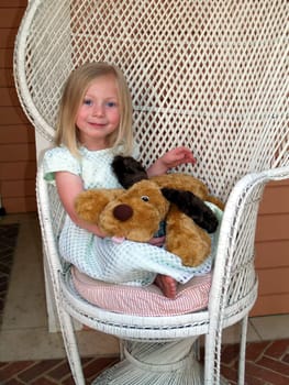 Young girl with stuffed puppy in her lap sitting in a rattan chair.