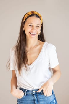 Portrait of confident beautiful woman with long brown hair, wearing casual clothes, standing in relaxed pose with hands in pockets, smiling with white teeth at camera, studio background
