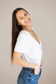 Portrait of confident beautiful woman with long brown hair, wearing casual clothes, standing in relaxed pose with hands in pockets, smiling with white teeth at camera, studio background