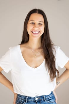 Portrait of confident beautiful woman with long brown hair, wearing casual clothes, standing in relaxed pose with hands in pockets, smiling with white teeth at camera, studio background