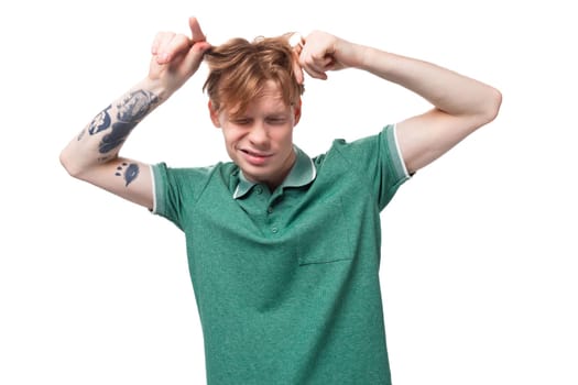 young stylish european guy with red hair with a tattoo on his forearm wears a green t-shirt touches his hair.