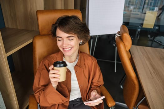 Portrait of happy young woman working in her office, sitting in chair with smartphone and coffee, drinking beverage from takeaway cup and smiling.