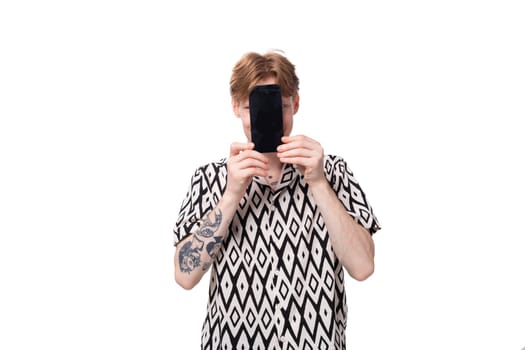 young caucasian man with red hair and a tattoo is dressed in a shirt with a rhombus pattern on a white background shows the screen of a smartphone with a mockup for advertising.