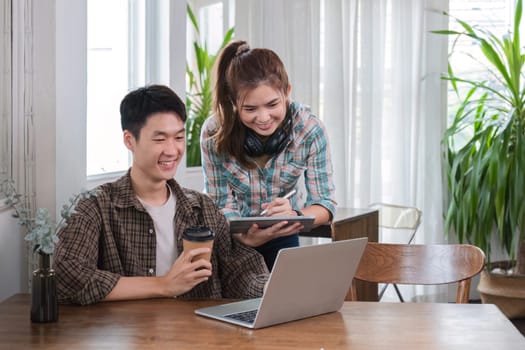 A happy young Asian man and a pretty girl are working on a laptop together, working on a co-project, sharing ideas and discussing work in a room..