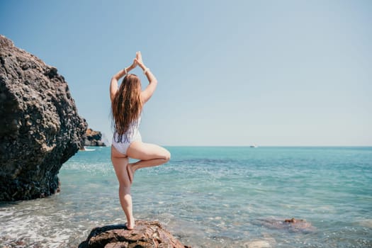 Woman sea yoga. Back view of free calm happy satisfied woman with long hair standing on top rock with yoga position against of sky by the sea. Healthy lifestyle outdoors in nature, fitness concept