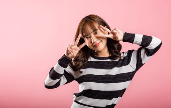 Asian happy portrait beautiful young woman smiling showing finger making v-sign victory gesture symbol near eye, studio shot isolated on pink background, female curly hair excited show two fingers