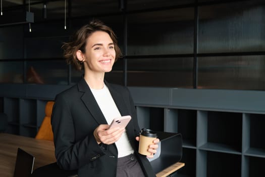 Portrait of beautiful saleswoman in suit, standing with coffee and smartphone, smiling, posing in conference room.
