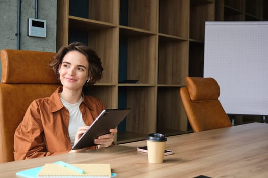 Portrait of happy young businesswoman, saleswoman in casual clothes, working in office, sitting with digital tablet and coffee, working on start-up.