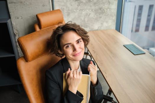 Top angle view of successful businesswoman, writing down information in notebook, working on planning her schedule, sitting in office and smiling at camera.