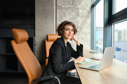 Image of corporate woman working in an office, sitting in front of laptop, preparing for business meeting, wearing black suit.