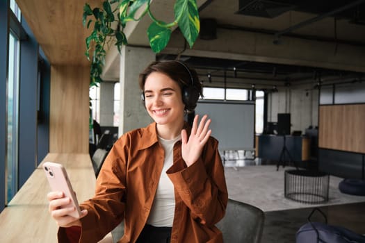 Portrait of young woman working in office, saleswoman says hello, video chats on smartphone app, connects to online meeting on mobile phone, wears headphones, works in co-working space.