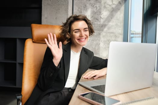 Cheerful businesswoman, office manager connects to video online meeting, saying hello and waving hand at laptop with friendly smiling face.