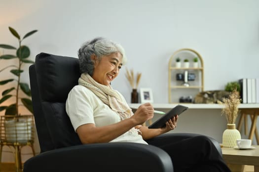 Positive elderly woman sitting on armchair and using digital tablet. People and technology concept.