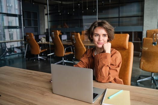 Portrait of beautiful working woman in an office, digital nomad entrepreneur sits at her workplace, does coding on computer, sitting in casual clothes.