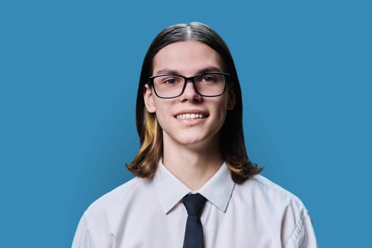 Headshot portrait of smiling teenage guy 18, 19 years old, on blue color studio background. Positive handsome young male in glasses shirt and tie looking at camera