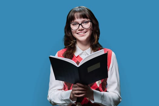 College university student reading exercise book, on blue color studio background. Smiling girl in glasses looking at camera, posing with notebook, education knowledge learning concept