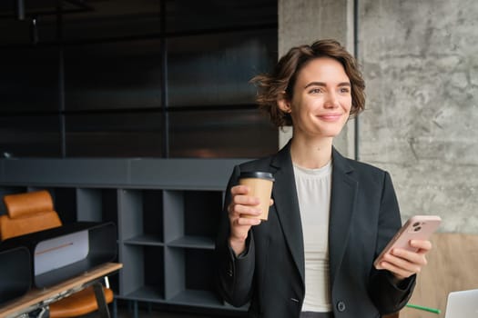 Portrait of successful businesswoman with her morning cup of coffee, standing near office window and smiling, reading the news on mobile phone app.