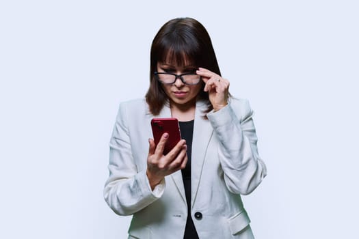 Portrait of surprised middle aged beautiful businesswoman with phone on white studio background. Serious mature female in suit, glasses looking at smartphone. Technology, business, people concept