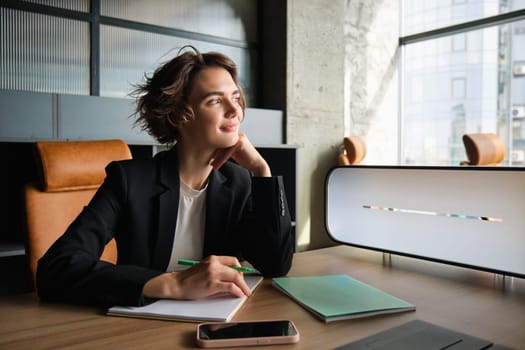 Portrait of young successful business woman, sitting in office with pen and papers, working on documents, waiting for meeting, preparing for corporate presentation.