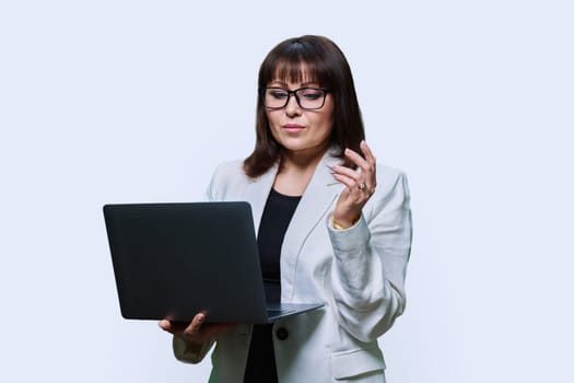 Mature business woman using laptop, on light studio background. Confident serious middle aged female in glasses suit looking at screen. Technology businessperson management teaching finance insurance