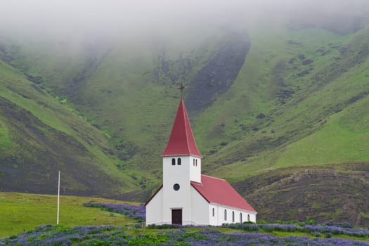 A little church in the south of Iceland surrounded by hills and green meadow on a foggy day.