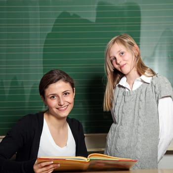 Cute teacher with textbook and pupil in front of the blackboard. Both smile at the camera