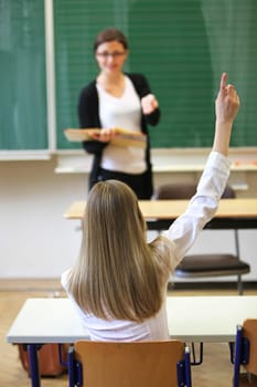 Elementary school student in elementary school pointing out. In the background,the teacher is standing in front of the blackboard with a book