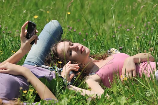 Two friends (teenage girls) lying on grass outside in nature - one girl is using cellphone and another sleeping