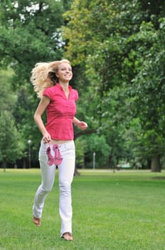 Smiling young woman running barefoot on grass in park and holding her shoes in hand